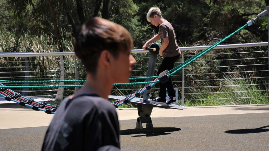 A wide shot showing a young boy standing on playground equipment with a woman in the foreground out of focus.