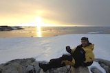 A bearded man in yellow and black outdoor clothing sits on a rock surrounded by ice in Antarctica.