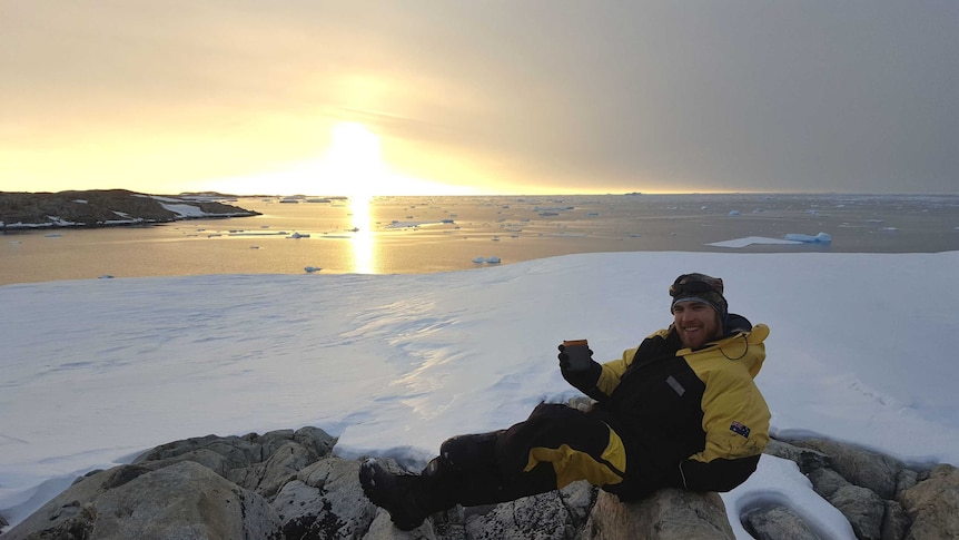 A bearded man in yellow and black outdoor clothing sits on a rock surrounded by ice in Antarctica.