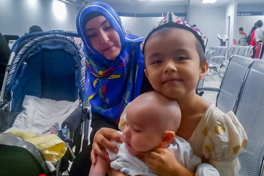 A girl wearing traditional Uyghur hat holds her baby brother in her lap with her mother looking on.