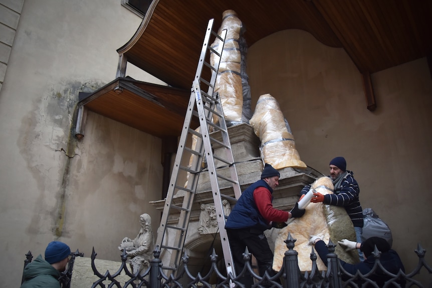 Two men on ladders using protective materials to wrap and tape up a statue.