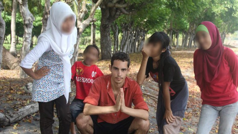A man wearing a red shirt squatting down while surrounded by four children.
