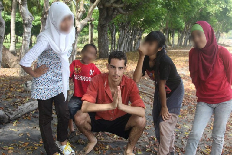 A man wearing a red shirt squatting down while surrounded by four children.