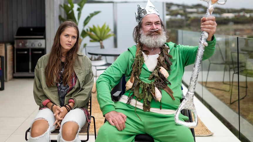A surly teen girl and an older man in a wheelchair, wearing a foil crown and holding a trident made from a spade, sit together
