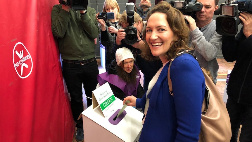 A woman laughs towards the camera as she votes, with media behind her.