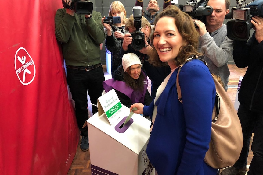 A woman laughs towards the camera as she votes, with media behind her.