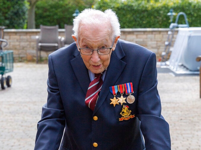 A man in a coat and war medals with white hair and glasses looks at the camera and walks with a walker.