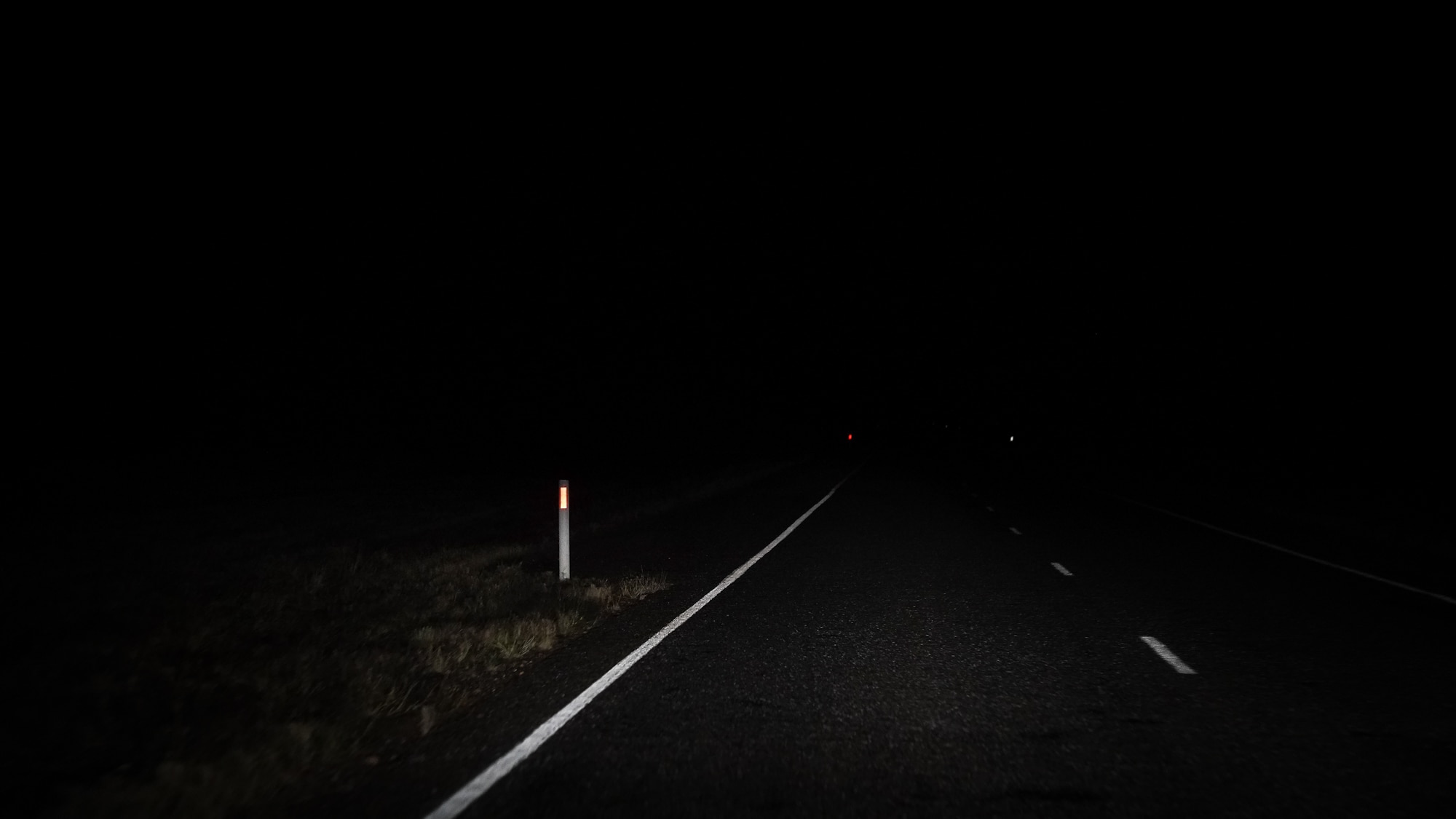 A dark lit image of the side of an outback road, with dim white light shining on the white paint lines and road marker pole.
