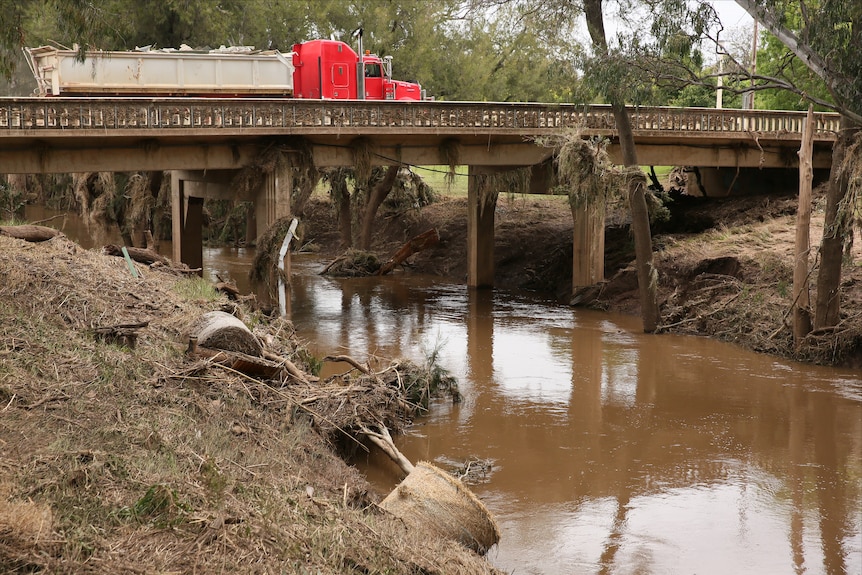 Debris including hay bales and canola is washed up against a stone bridge over a brown creek.