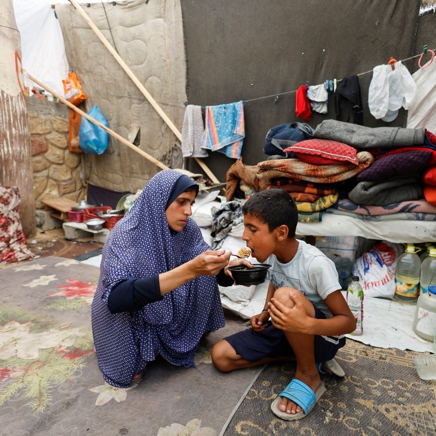 A woman and a young boy crouch on the ground. The woman is helping the boy eat. They are looking at each other.