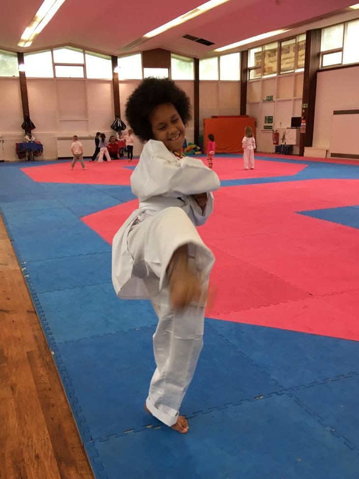 Une jeune fille avec un grand afro brun dans une tenue de taekwondo blanche sourit en donnant un coup de pied à la caméra