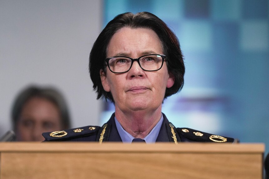 A woman with short dark hair and glasses in a police uniform stands at a podium.