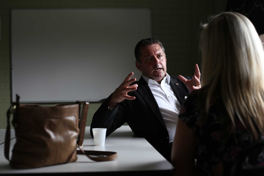 A man leaning against a table in an office talking to a blonde woman.