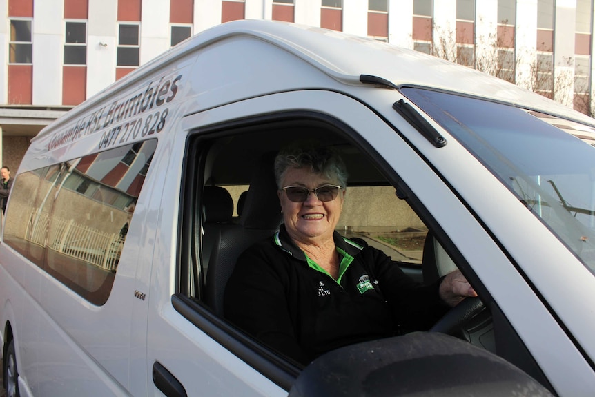 A woman in sunglasses smiles widely from behind the wheel of a bus, with the RSL building behind her