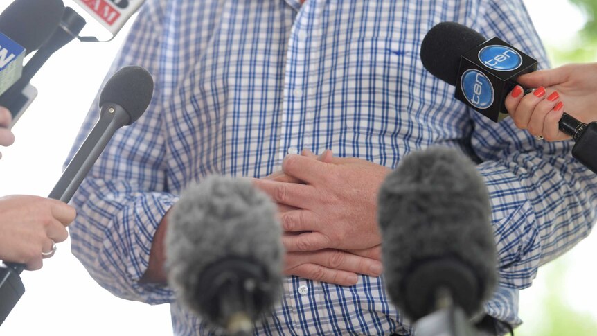 Barnaby Joyce's hands are held across his stomach, in front there are several microphones
