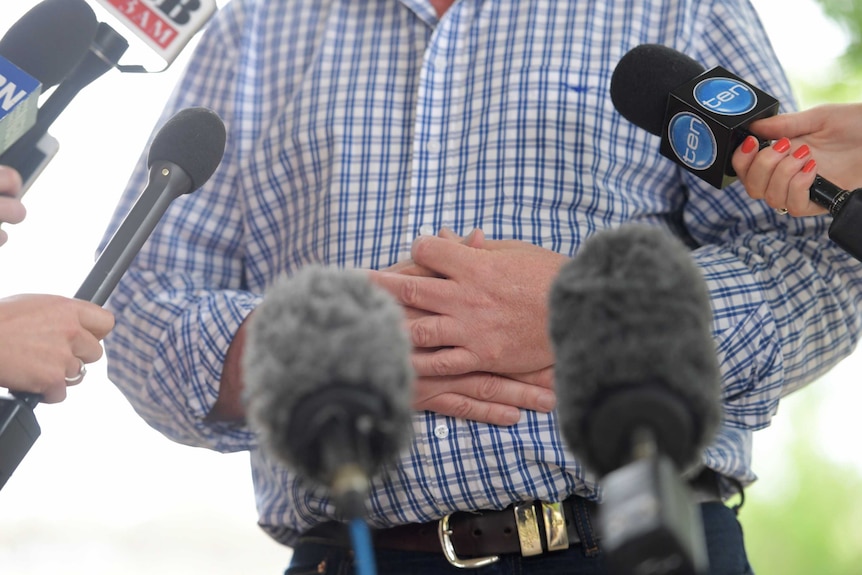 Barnaby Joyce's hands are held across his stomach, in front there are several microphones