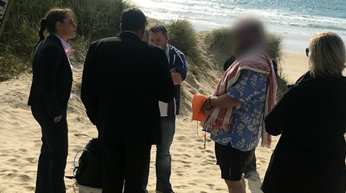 Five people gathered on a beach, man second from right has his face blurred.