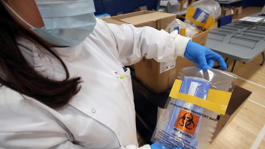 a woman wearing a face mask and protective gear holds a pack holding a testing kit