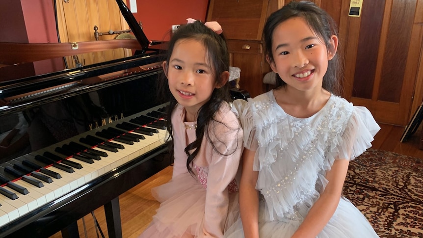 two girls, one in a pink dress and one in a blue dress, are smiling at the camera sitting at a piano
