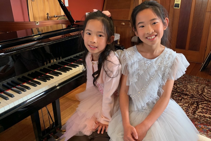 two girls, one in a pink dress and one in a blue dress, are smiling at the camera sitting at a piano