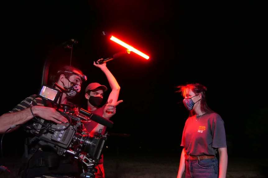 A young man with a professional camera and a young man holding a red light have a discussion with a young woman at nighttime.