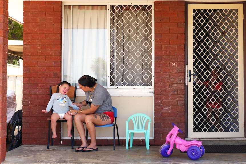 Mère et jeune fils sont assis sur la terrasse d’une maison en briques sombres.  Chaise pour enfants et jouet rose présents. 