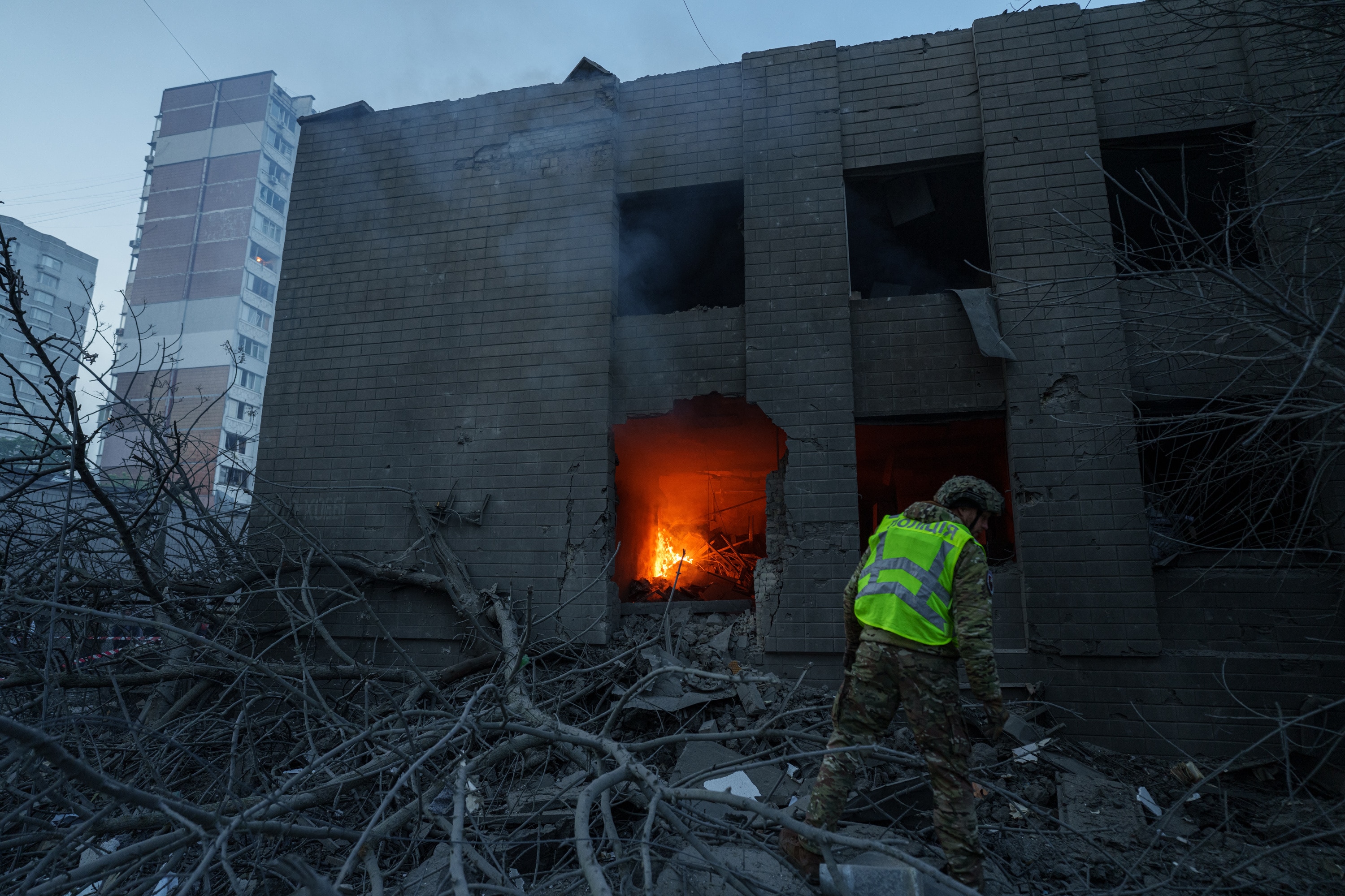 A fire burning through the blown out windows of a damaged building as a police officer walks through debris outside.