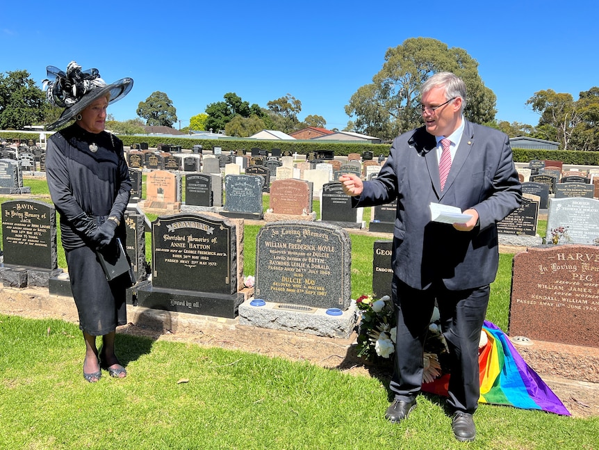 A man wearing a suit and tie and a drag queen wearing a large black hat and dress in a cemetery