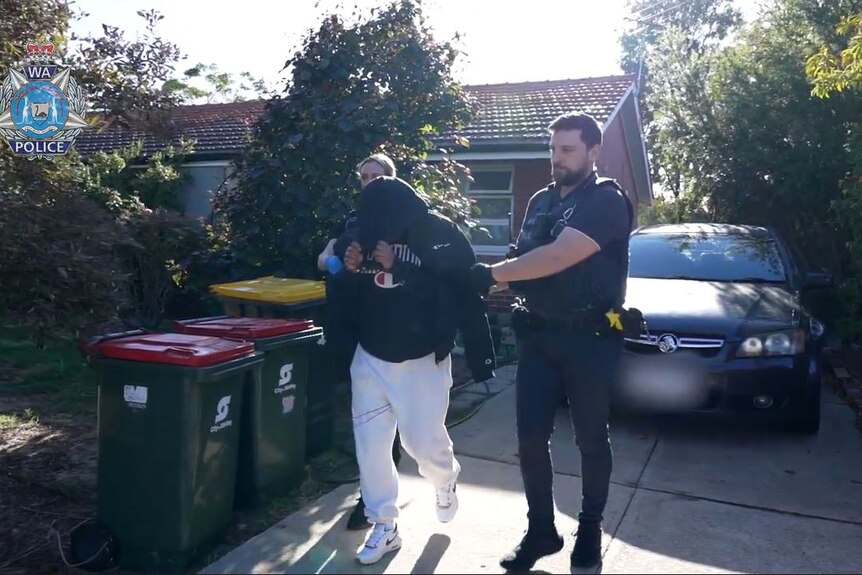 A man with a hoddie over his face is led out of a house by two police officers.