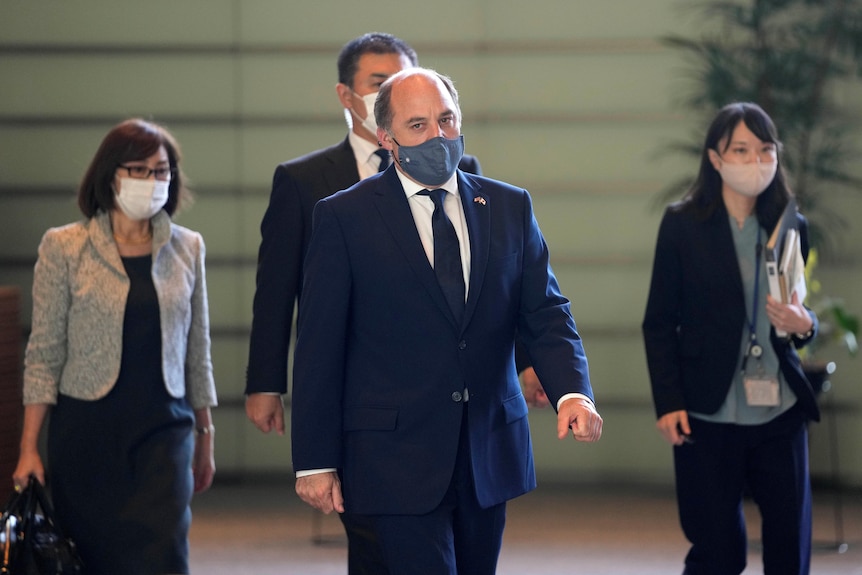 A middle-aged balding man in a dark suit and face mask strides through a room flanked by two Asian women in masks