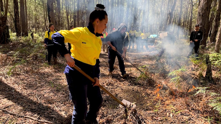 Man and woman with rake-hoes putting out a fire in leaf litter as others watch on