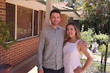 A couple stand in front of a house smiling