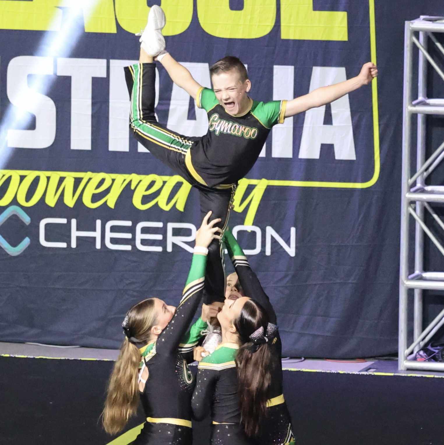 Young boy in team lycra green and black standing held up by girls holding one leg up behind him as flier in a cheer stunt.