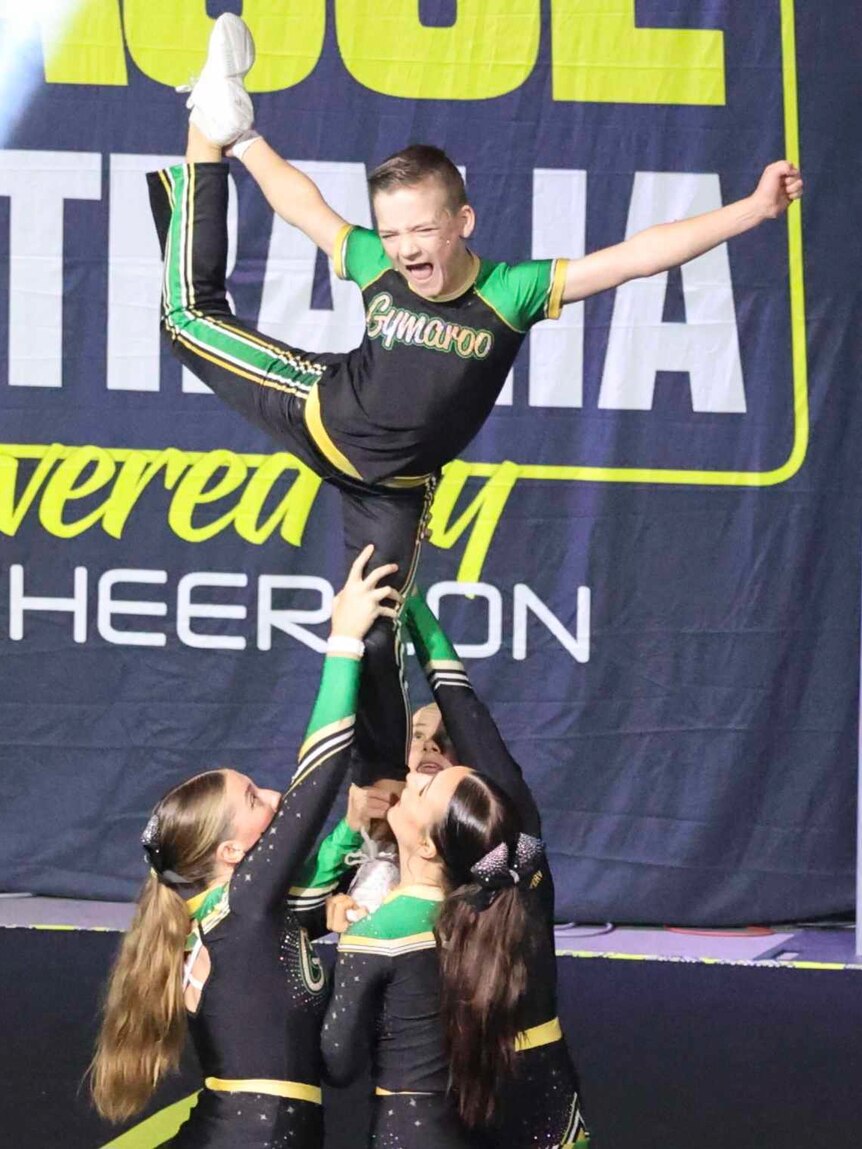 Young boy in team lycra green and black standing held up by girls holding one leg up behind him as flier in a cheer stunt.