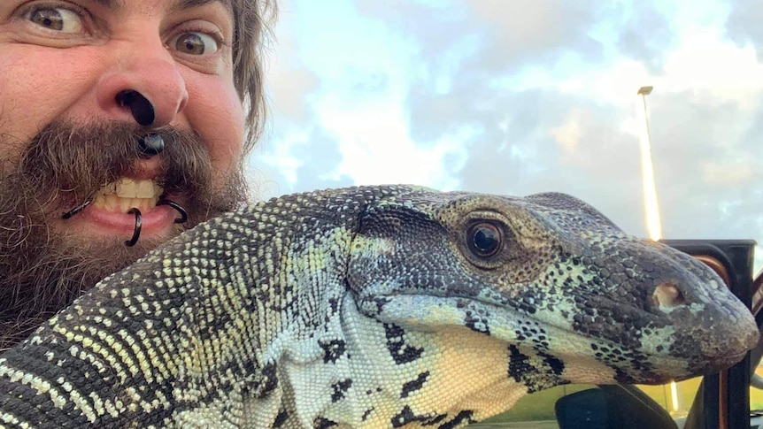 A bearded man with several face piercings holds up his pet lace monitor.