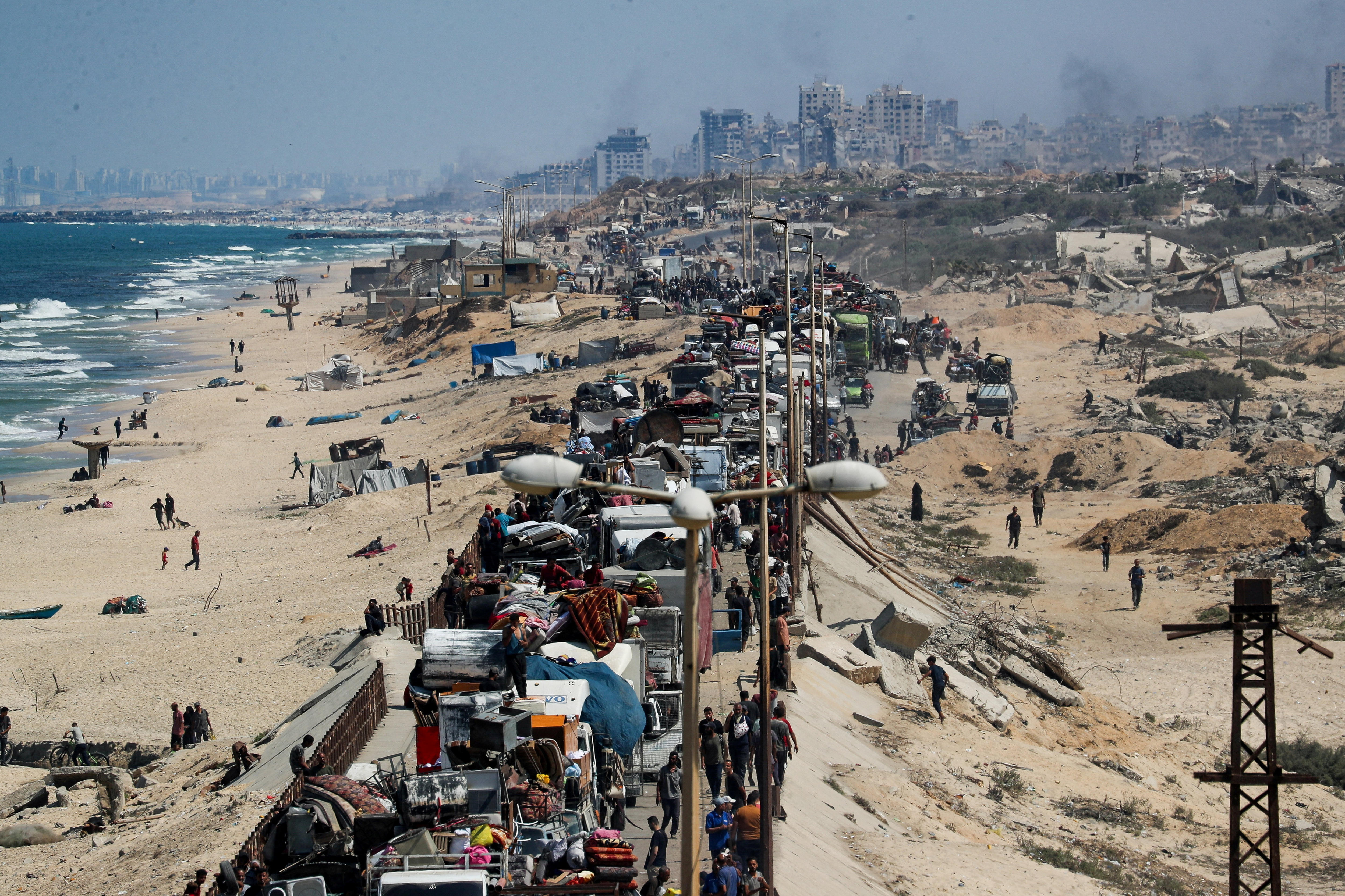 A crowd of people and vehicles squished on a thin road with beach sand on either side, in the distance destroyed buildings