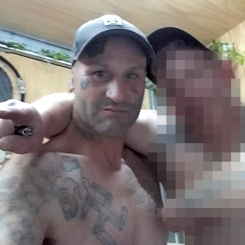 A man with a Swastika tattoo on his chest wearing a baseball cap but no shirt looks at the camera.