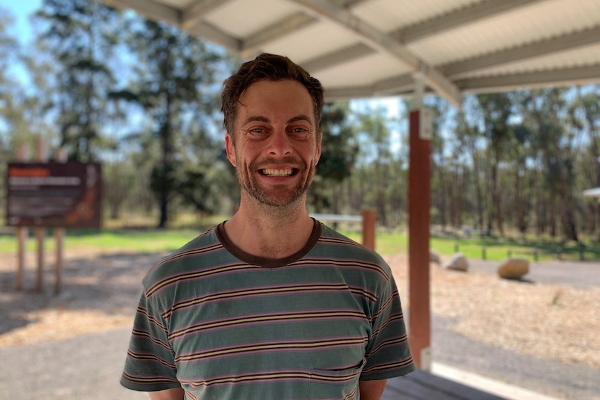 A smiling man in his 30s with facial stubble standing in a park picnic area.