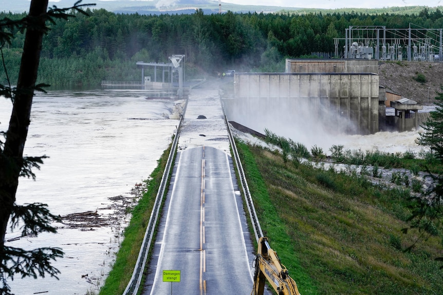 A bridge going across a dam being overflown with water with surrounding trees and grass being covered