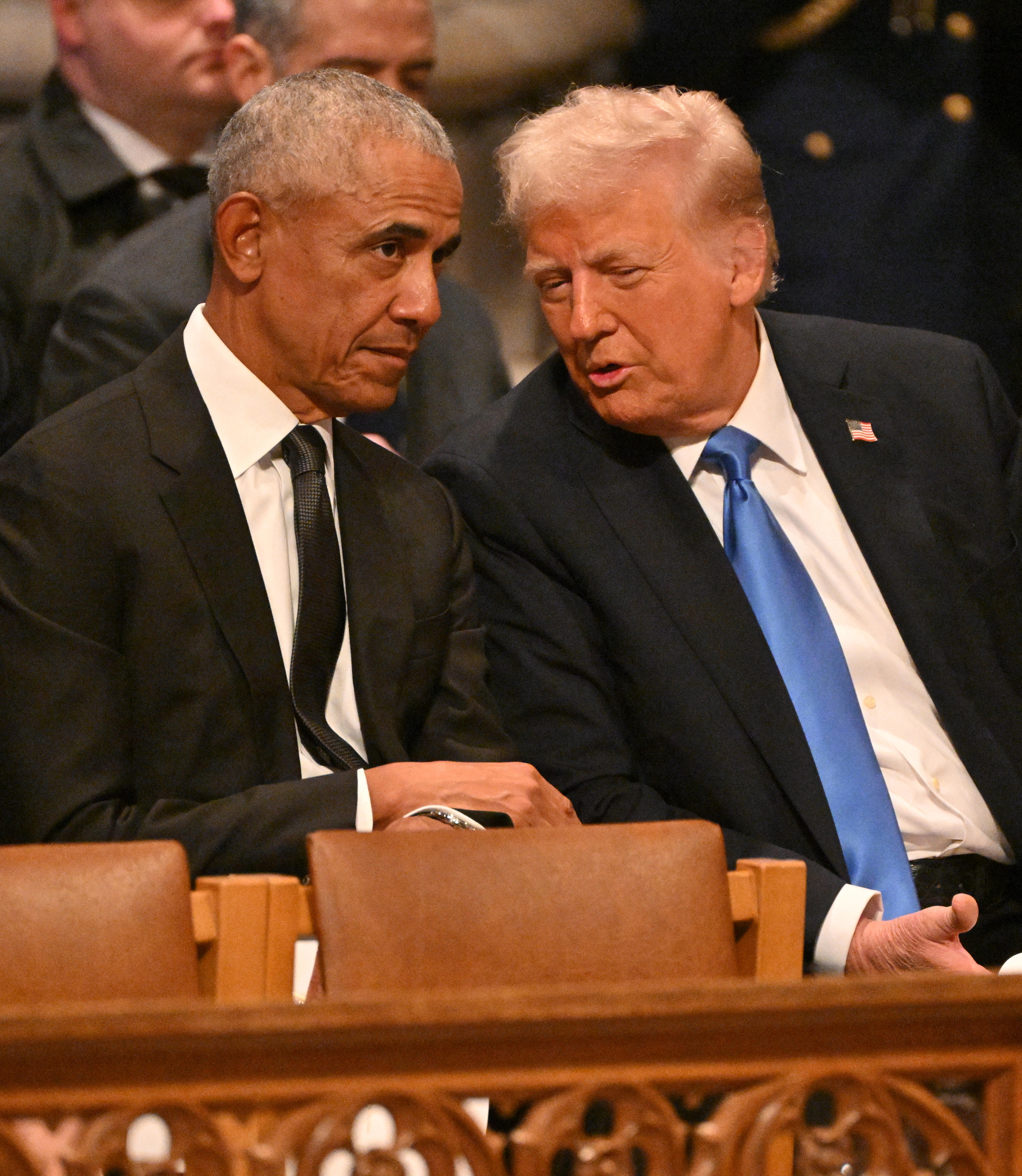 Barack Obama and Donald Trump, both dressed in dark suits, sit next to each other at a funeral.