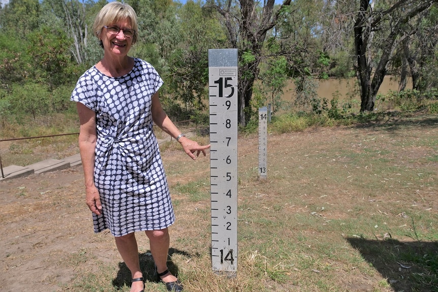 Anne Chater standing beside a flood gauge, pointing at the 2010 peak level of 14.7 metres, stairs, grass, trees, murky river.