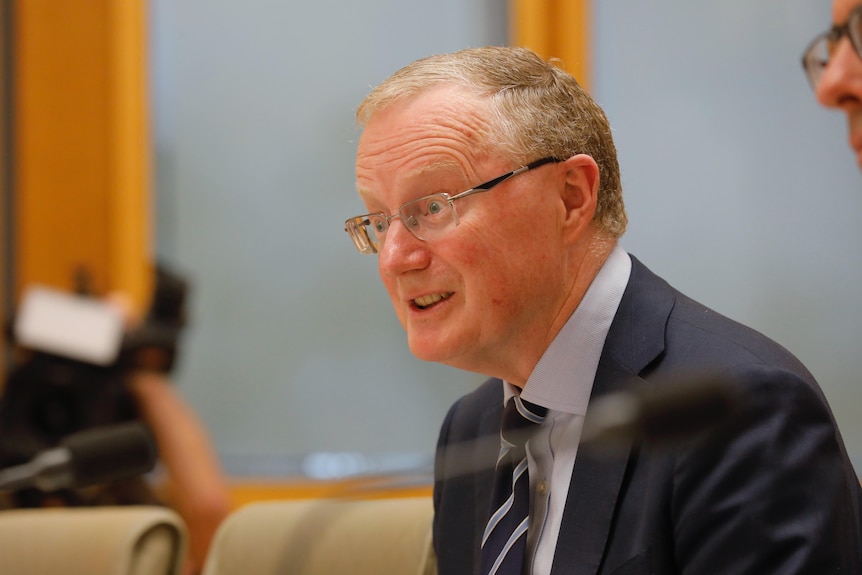 Philip Lowe wearing glasses and a suit sits at a table and speaks.