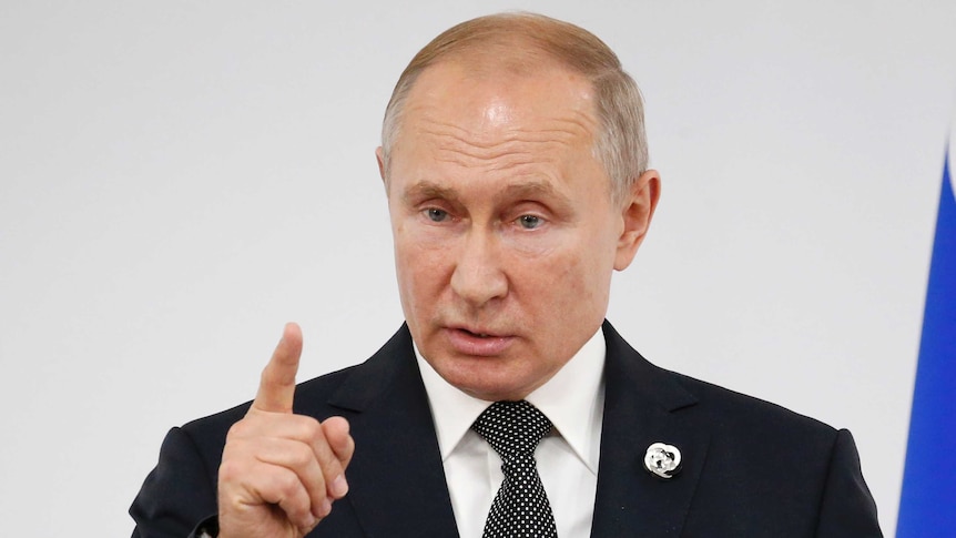 Vladimir Putin points a finger upwards while speaking into two microphones.