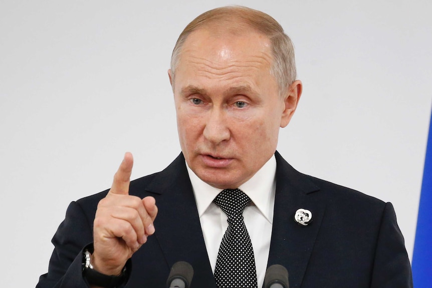 Vladimir Putin points a finger upwards while speaking into two microphones.