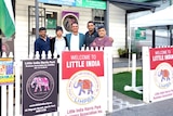 A group of men in the front yard of a house with signs on the fence saying 'Welcome to Little India'