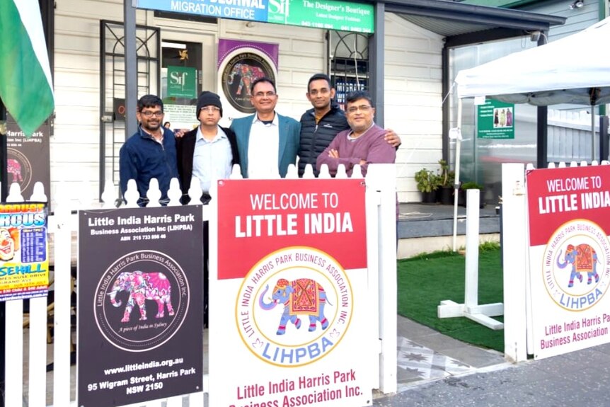 A group of men in the front yard of a house with signs on the fence saying 'Welcome to Little India'