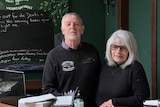 A man and a woman stand behind a desk and look at the camera.