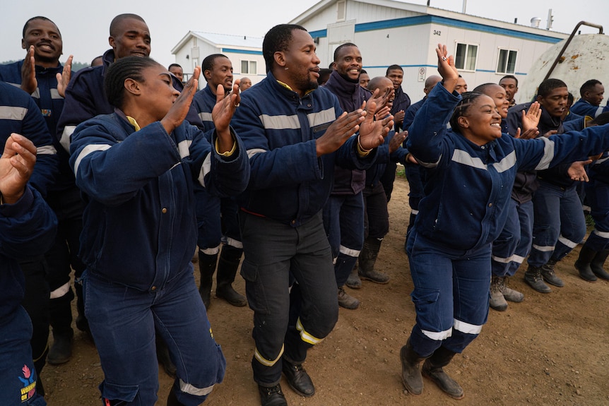 A group of South African firefighters in navy uniforms are captured mid-dance, some clapping, some with their hands in the air.