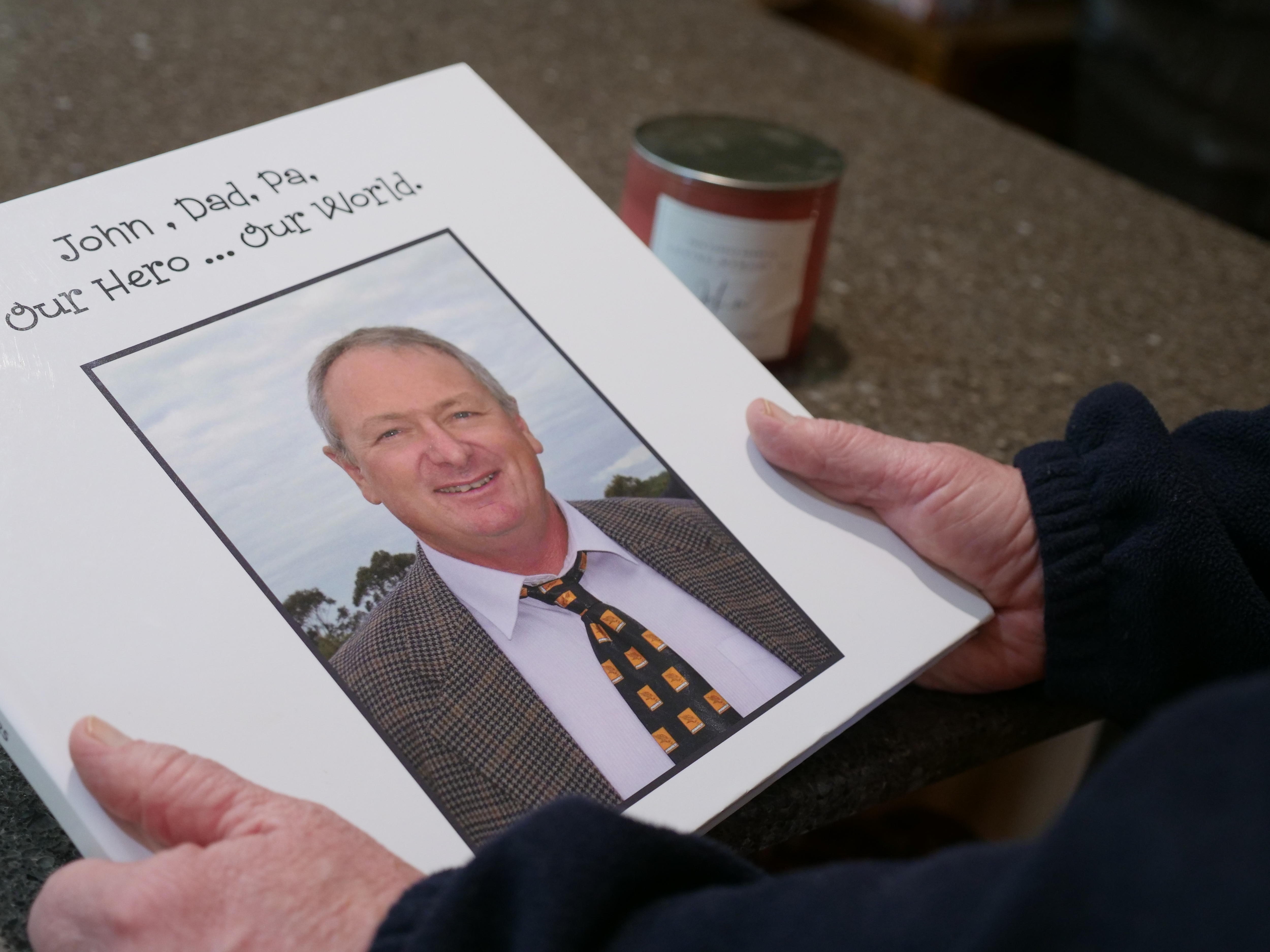 A woman holds a photo memory book with the words 'John, Dad, Pa, Our Hero ... Our World'.