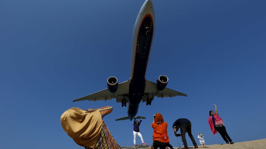 People crouch and pose on the sand taking photos and posing as a plane looms in the blue sky overhead